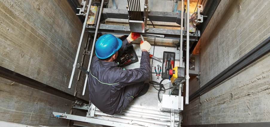 View from the top of an elevator shaft where a technician is working on the machinery below.