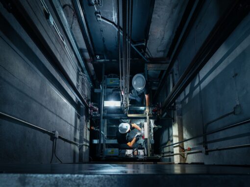 View from the top of an elevator shaft where two technicians are working on the machinery below.