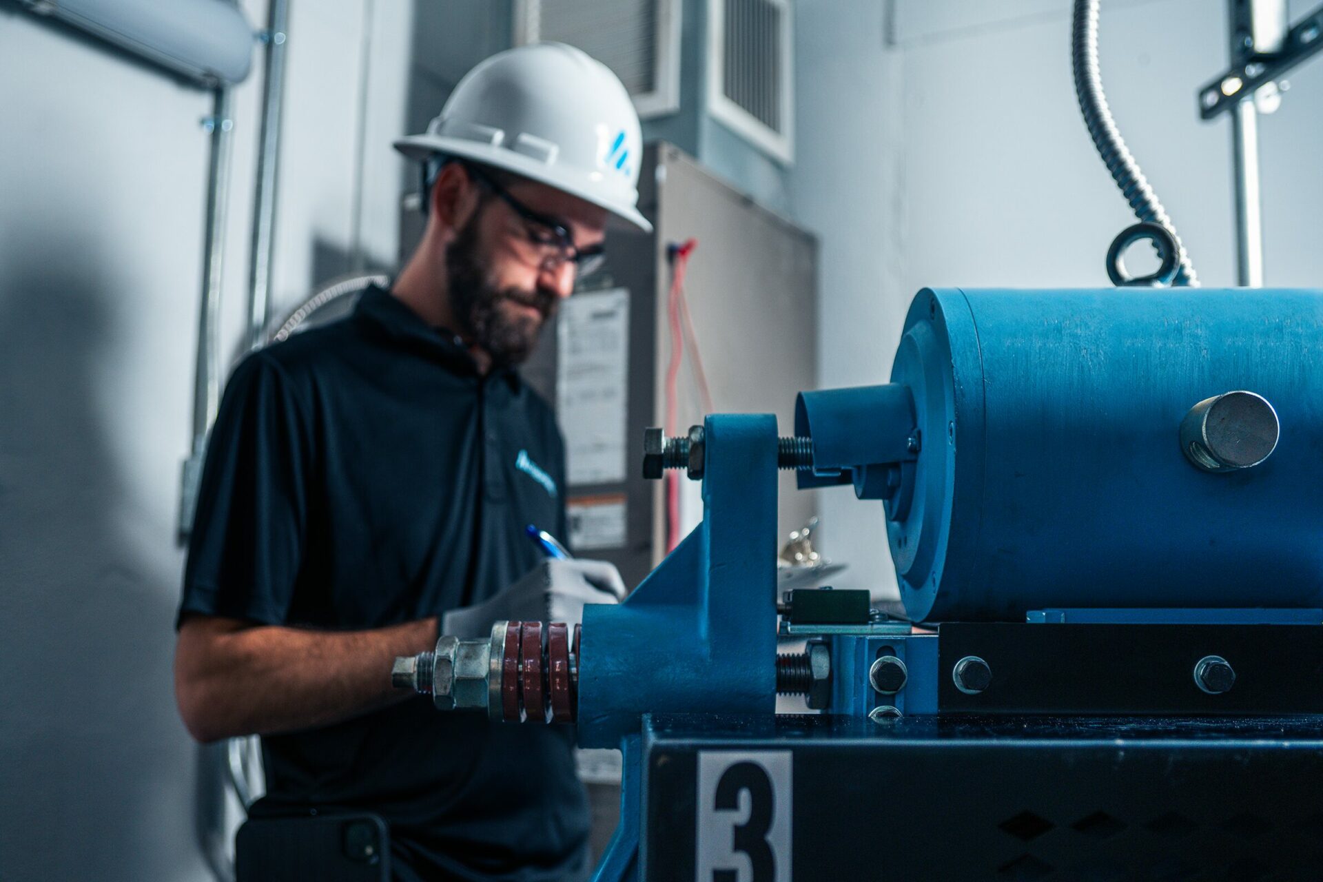 An elevator technician with a helmet writing on a clipboard beside a blue elevator motor.