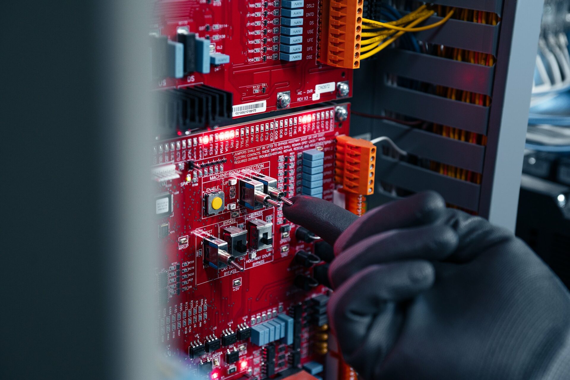 A technician's hand adjusting components on a red circuit board within an elevator system.