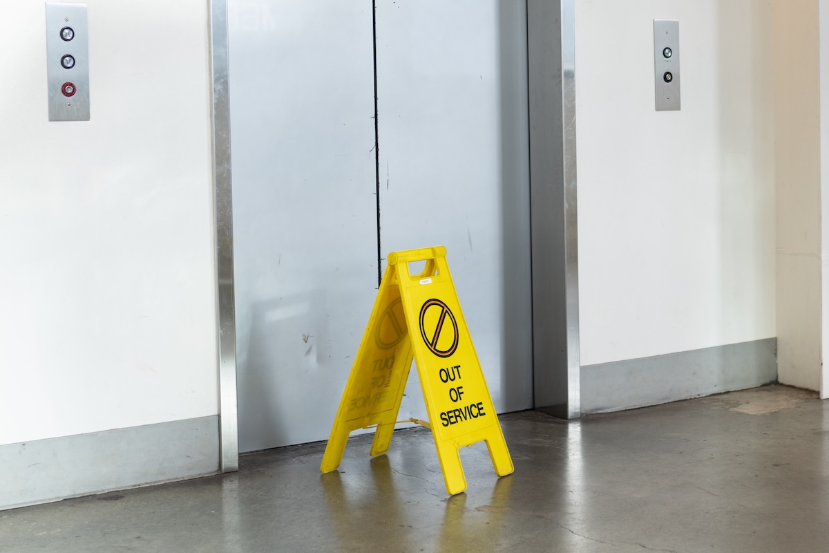 A yellow 'Out of Service' sign in front of a closed elevator door in a building lobby.