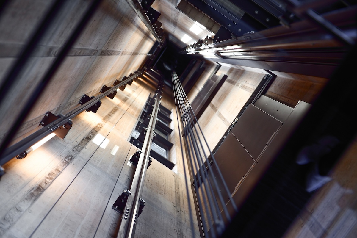 View looking down a deep elevator shaft from the top showing multiple cables and the top of the elevator car.