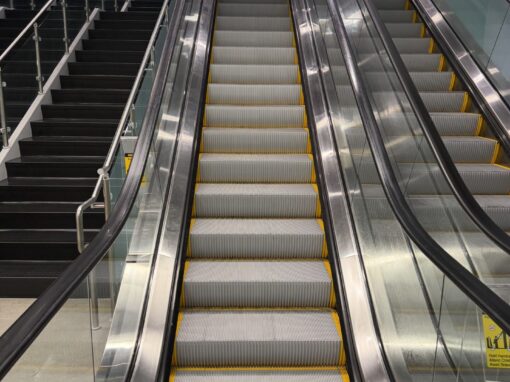 Indoor escalators and adjacent staircases in a commercial building, shown from the base looking upward toward the upper level.