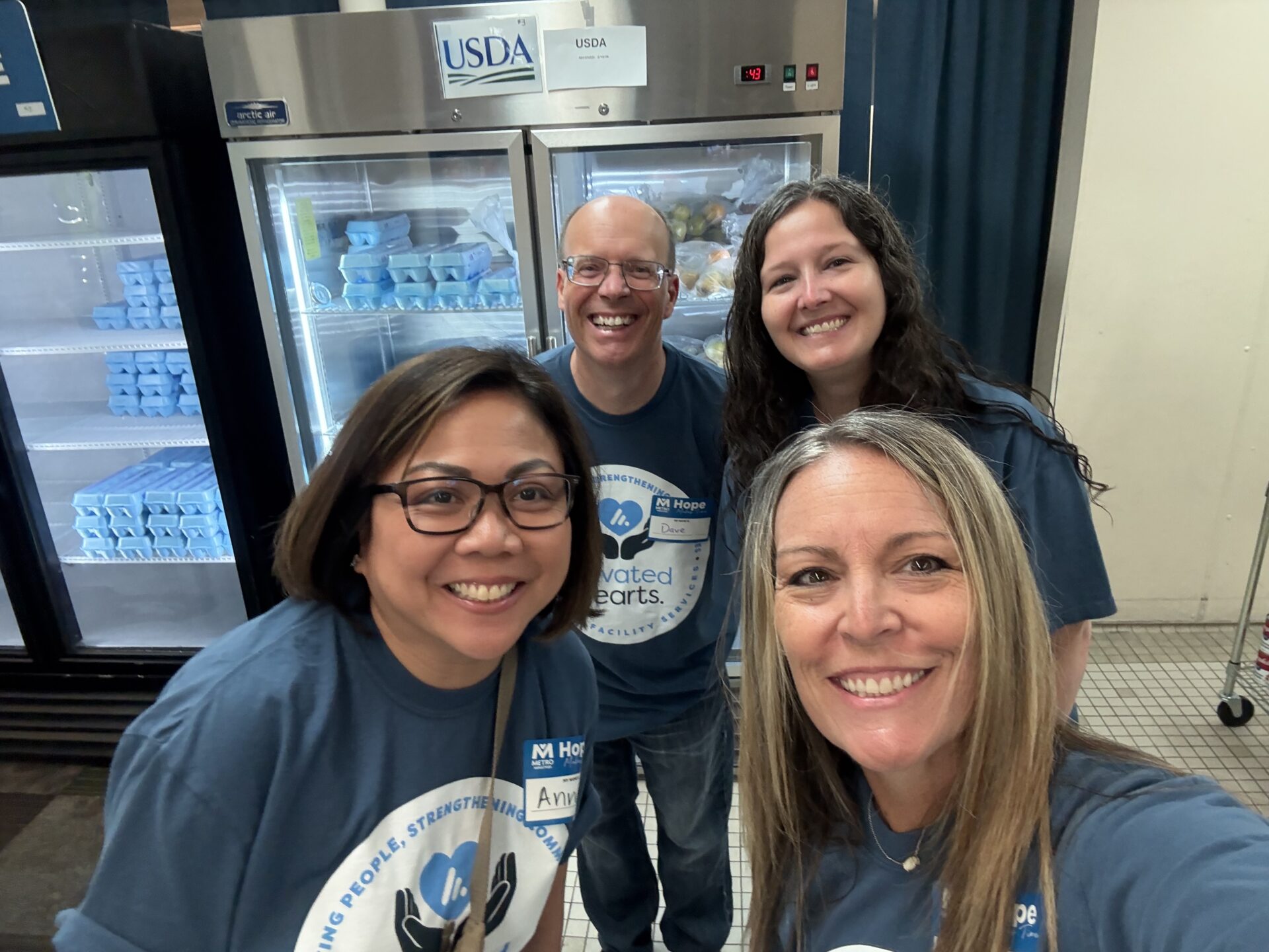 Elevated Facility Services employees pose for a group image inside a Metropolitan Ministries food pantry, with refrigerated food items visible behind them.