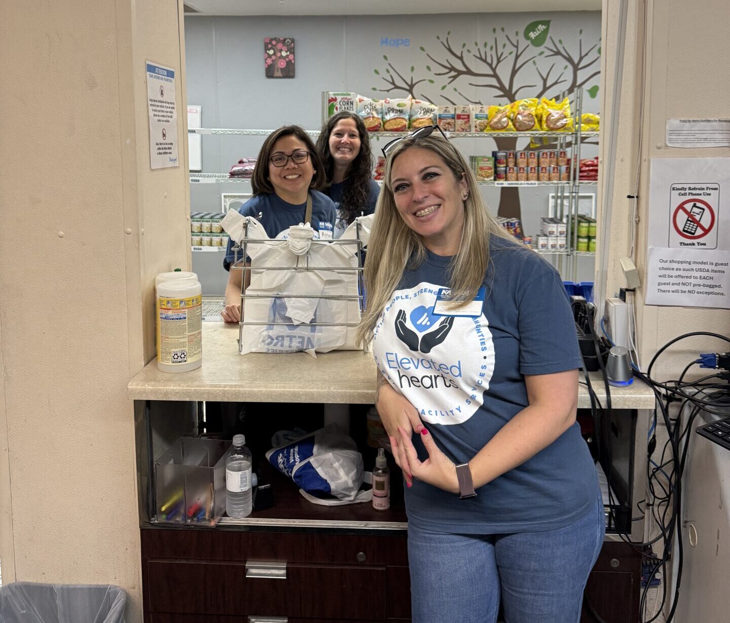 Elevated Facility Services employees pose at a food pantry station inside Metropolitan Ministries, with shelves of packaged food visible in the background.
